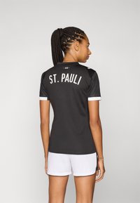 Zwarte voetbalshirt met "ST. PAULI" en het jaar "1910" gedrukt op de rug, witte accenten op de mouwen, gecombineerd met witte korte broek.