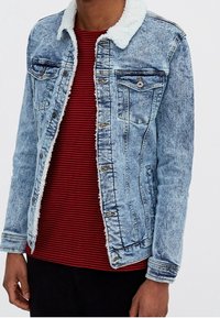 Hombre vistiendo una chaqueta de mezclilla azul clara con cuello de forro polar blanco sobre una camiseta a rayas rojas y negras y pantalones negros, de pie con los brazos relajados.