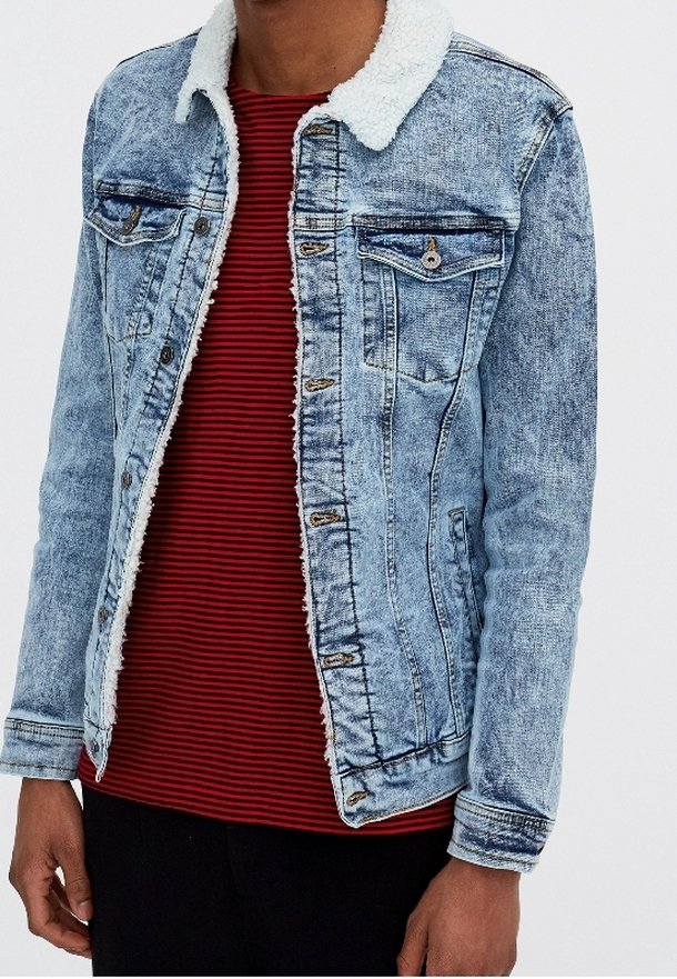 Hombre vistiendo una chaqueta de mezclilla azul clara con cuello de forro polar blanco sobre una camiseta a rayas rojas y negras y pantalones negros, de pie con los brazos relajados.