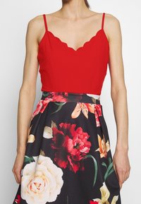 Donna che indossa un top senza maniche rosso con bordi a scallop, abbinato a una gonna nera decorata con grandi stampe floreali rosse, bianche e arancioni.