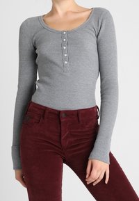 Grå vadderad långärmad topp med rundad halsringning och femknapps knäppning, tillsammans med högmidjade burgundy byxor.