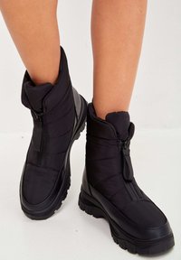 Botas de tobillo aisladas en negro con un diseño acolchado, suela de goma y un cierre lateral con una lengüeta de tela. Parte superior texturizada para proporcionar Calidez.