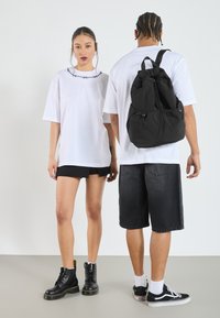 Svart ryggsäck med dragkedjor, bärs av en person i en vit T-shirt. Ljust grå sneakers och svarta shorts syns också. Svarta kängor står till vänster.