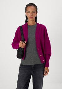 Cardigan lavorato a maglia magenta con una vestibilità comoda, maniche a coste e tre bottoni color oro. Indossato sopra una maglietta grigia e abbinato a jeans scuri.