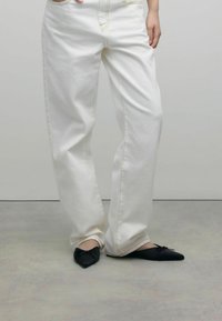 Jean blanc à jambes larges avec des coutures jaunes, associé à des ballerines noires pointues avec un détail en nœud à l'avant. Textures lisses.