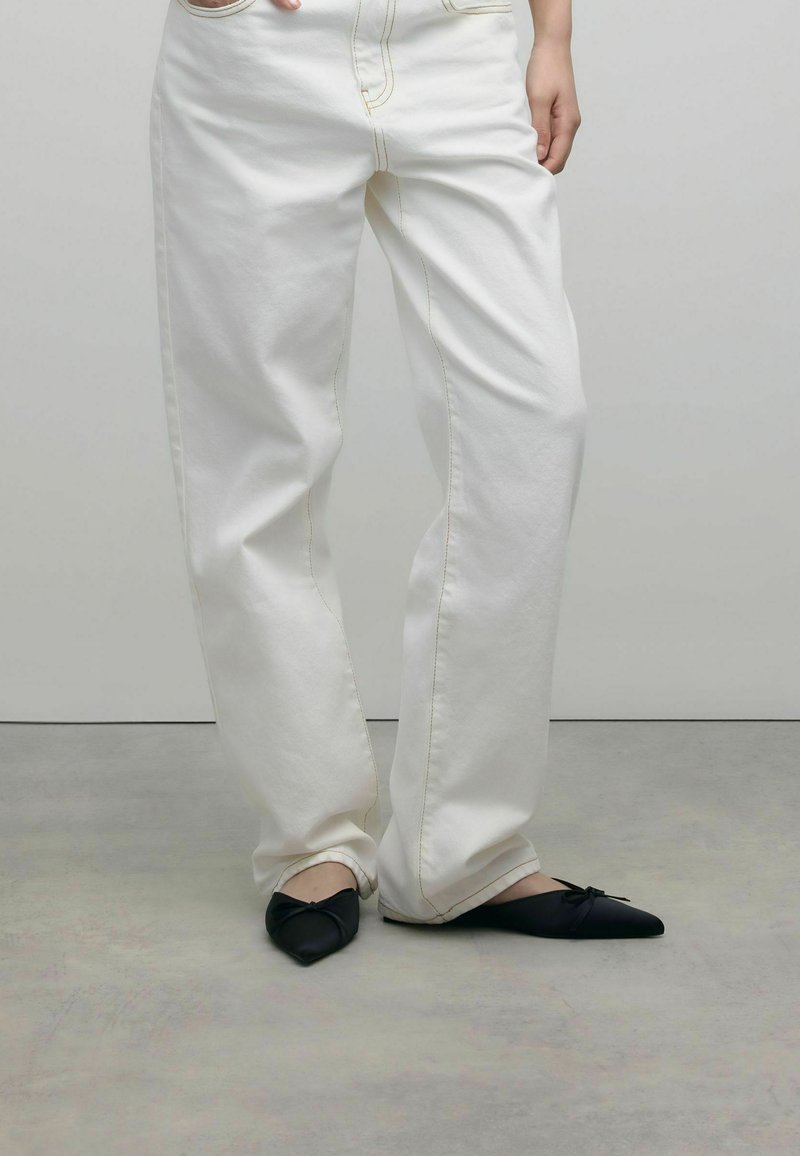 Jean blanc à jambes larges avec des coutures jaunes, associé à des ballerines noires pointues avec un détail en nœud à l'avant. Textures lisses.