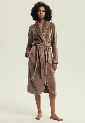 Kobieta z kręconymi włosami stoi boso, nosząc miękki, średniej długości brązowy szlafrok związywany w talii, na jednolitym tle.