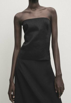 Femme portant un haut noir sans bretelles et une jupe noire, debout devant un fond uni gris clair.
