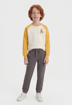 Παιδί με κόκκινα μαλλιά στέκεται φορώντας λευκό και κίτρινο μακρυμάνικο πουκάμισο με σχέδιο, γκρι παντελόνι με κορδόνι και λευκά αθλητικά παπούτσια σε φωτεινό φόντο.