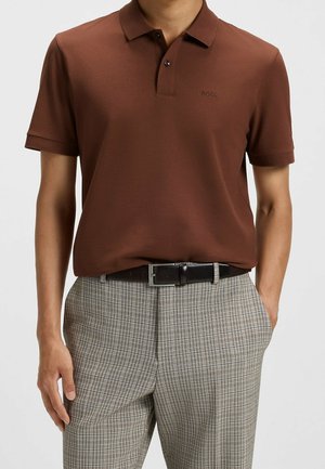 Mann trägt ein braunes kurzärmeliges Poloshirt, das in graue karierte Hose mit schwarzem Ledergürtel gesteckt ist.