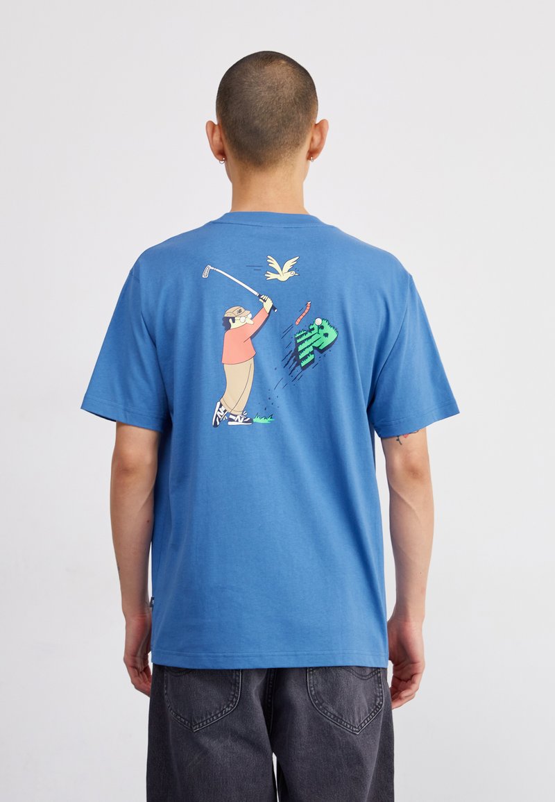 Zila kokvilnas T-kreklā ar īsām piedurknēm. Grafika uz muguras attēlo cilvēku golfa spēlē, virs kura lido putns, un ir redzami zaļie zāles akcenti.