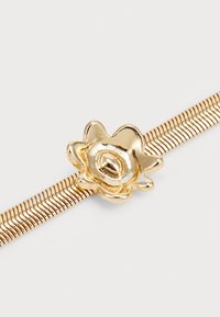 Broche en forma de flor de oro con un acabado brillante, montada sobre una banda metálica retorcida con estrías texturizadas. Superficie lisa con pétalos detallados.