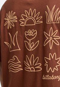 Braunfarbiges Baumwoll-T-Shirt mit verschiedenen beige gestickten Designs von Blumen, Wellen und Gras sowie dem Markentext "billabong" am unteren Rand.