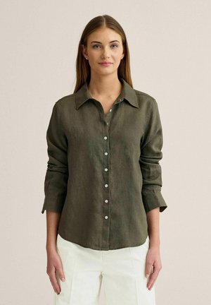 Ung kvinde med langt, glat hår iført en olivengrøn skjorte med knapper og hvide bukser, stående foran en ensfarvet baggrund.