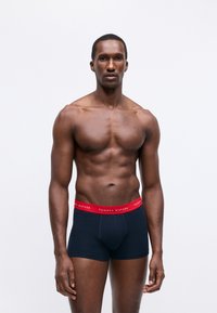 Muskuļots vīrietis stāv pret baltu fona, ģērbies tumši zilās Tommy Hilfiger bokseru biksēs ar sarkanu jostu.
