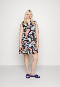 Vestido floral com base azul marinho e flores multicoloridas, design sem mangas e decote em V; combinado com sandálias roxas de tiras.