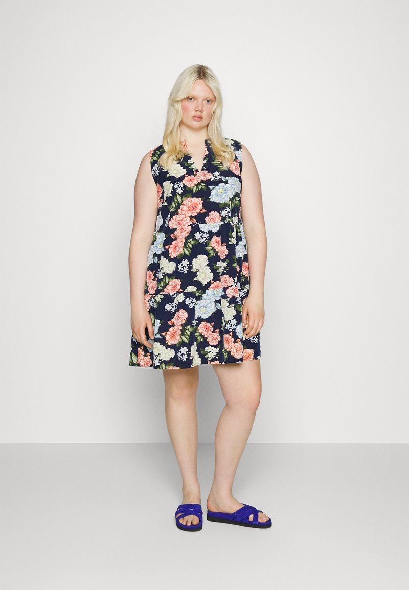 Vestido floral com base azul marinho e flores multicoloridas, design sem mangas e decote em V; combinado com sandálias roxas de tiras.