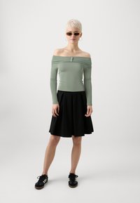 Off-the-shoulder, ljusgrön långärmad topp kombinerad med en svart veckad kjol och svarta sneakers med vita ränder. Minimalistisk design, figurnära passform.