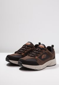 Brązowe buty sportowe wykonane z mieszaniny zamszu i siatki, z czarnymi akcentami, teksturowanym wzorem i pomarańczowym logo na języku.