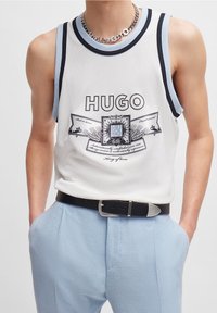 Débardeur en maille blanche avec des bordures noires et bleu clair, arborant un logo graphique central. Associé à un pantalon bleu clair, maintenu par une ceinture noire.