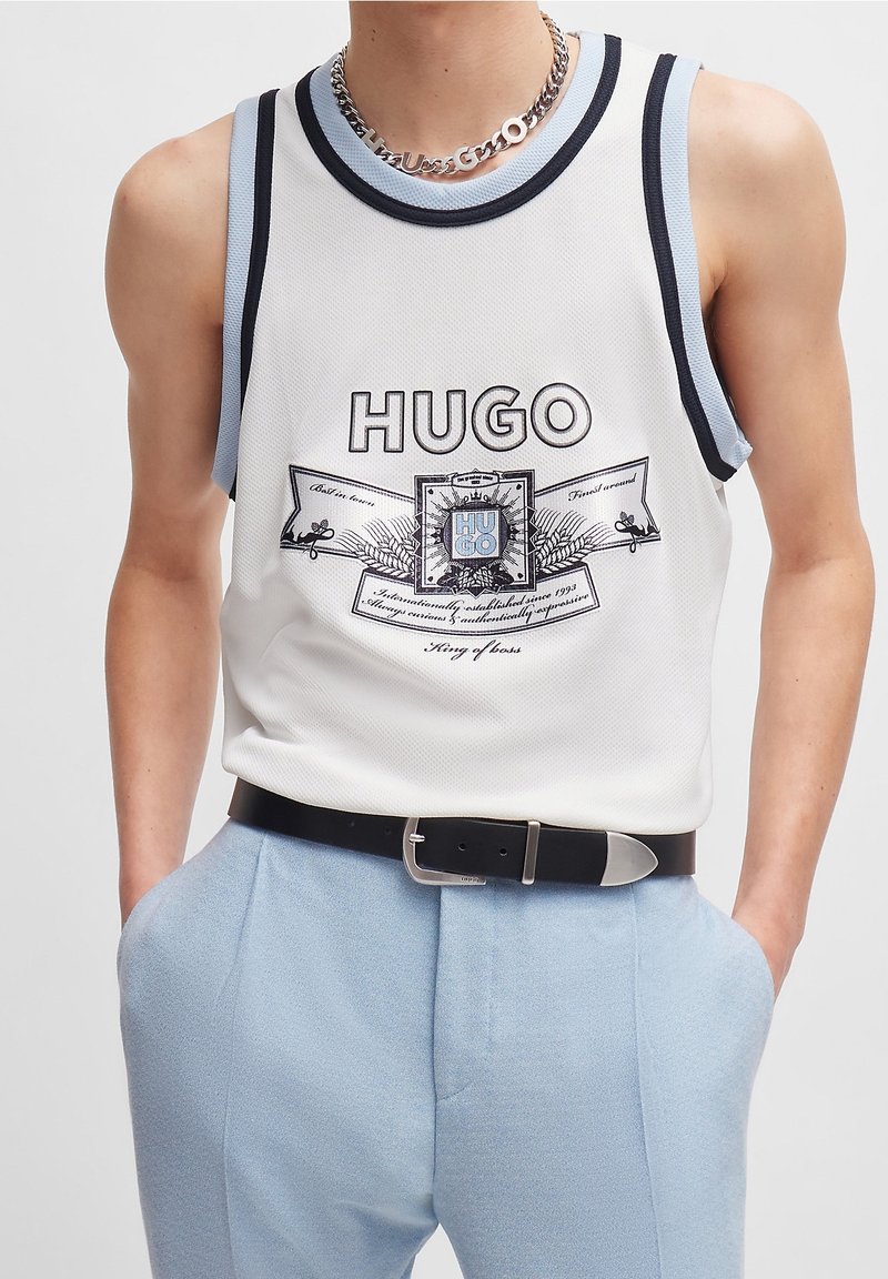 Débardeur en maille blanche avec des bordures noires et bleu clair, arborant un logo graphique central. Associé à un pantalon bleu clair, maintenu par une ceinture noire.