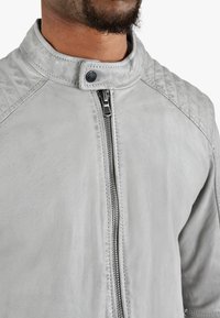 Uomo che indossa una giacca di pelle grigia chiara con dettagli trapuntati sulle spalle e chiusura a zip, abbottonata al collo con un bottone a scatto.
