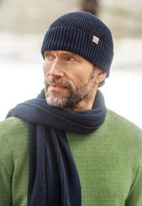 Navy ribbed knit beanie and scarf on a person wearing a green textured sweater, outdoors with a blurred snowy background.