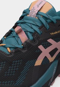 Κοντινή λήψη μπλε-πράσινου και μαύρου παπουτσιού πεζοπορίας Asics με ροζ λογότυπο, κορδόνια με σχέδιο και τις λέξεις "TRABUCCO" και "FF BLASTMAX" στο πλευρό.