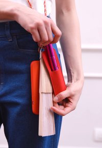 Porta bolígrafos de cuero rojo con un bolígrafo morado, una funda de teléfono naranja y un pequeño libro sostenido en una mano, sobre un fondo claro.