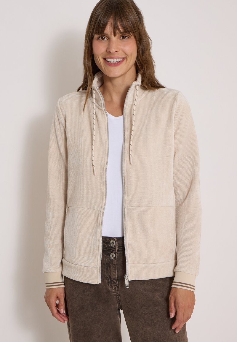 Femme souriante portant une veste beige zippée sur une chemise blanche et un pantalon brun foncé, debout devant un fond clair uni.