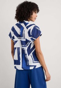 Top a maniche corte in tessuto bianco con un audace motivo astratto blu. La camicia ha una vestibilità rilassata con colletto classico e orlo leggermente curvo.