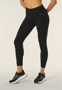 Leggings neri a vita alta realizzati in tessuto liscio. Presentano un logo discreto sul lato e un design aderente, abbinati a sneakers nere texturizzate.