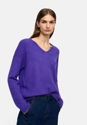 Fioletowy sweter z dekoltem w serek, wykonany z miękkiego, teksturowanego materiału, o luźnym kroju i długich rękawach, zestawiony z ciemnymi dżinsami. Prosty design, bez widocznych grafik.