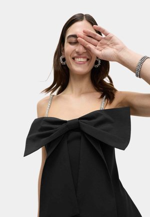 Vestido negro con un gran lazo en la parte delantera, con tirantes brillantes y una textura suave. Modelo mostrando una pose juguetona con la mano levantada.