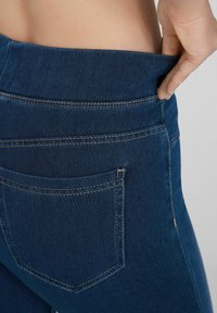 Jeans elásticos de un azul oscuro con una textura suave, que presentan una cinturilla ancha y un bolsillo trasero con costuras en contraste.