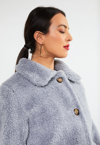Grå, fluffy, tekstureret frakke med krave og to knapper. Guld trekantede øreringe synlige. Minimalistisk design med fokus på komfort.