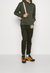 Oliwkowa bluza z kapturem ze ściągaczami, czarne spodnie typu cargo z kieszeniami oraz jaskrawo pomarańczowo-żółto-czarne buty sportowe. Beżowa torba na ramię.