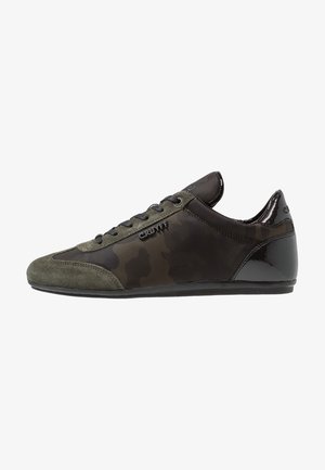 Baskets basses pour hommes avec un motif camouflage noir et vert foncé, embout en daim, talon noir brillant, et logo "Cruyff" sur le côté.