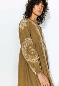 Robe vert olive avec des motifs blancs brodés le long des manches et de la poitrine, avec un devant à boutons et une texture froncée sous la poitrine.