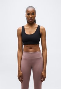 Γυναίκα που στέκεται με το πρόσωπο προς τα εμπρός, φορώντας μαύρο αθλητικό σουτιέν και μωβ ψηλόμεσο κολάν, μπροστά σε απλό φωτεινό φόντο.