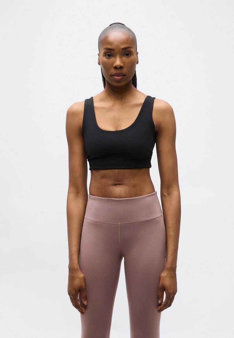 Γυναίκα που στέκεται με το πρόσωπο προς τα εμπρός, φορώντας μαύρο αθλητικό σουτιέν και μωβ ψηλόμεσο κολάν, μπροστά σε απλό φωτεινό φόντο.