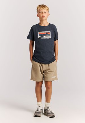 Marineblaues T-Shirt mit einem Grafikdruck in Beige und Orange, kombiniert mit beigen Shorts und grauen Sneakers mit weißen Schnürsenkeln sowie gestreiften Socken.