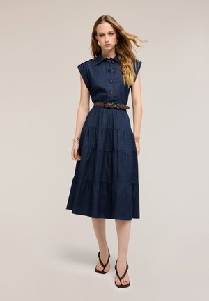 Kvinde iført en ærmeløs mørk denim midi-kjole med knapper, et brunt bælte, armbånd og sorte sandaler med firkantet tå, stående foran en neutral baggrund.