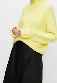 Pull jaune tricoté avec un col montant, doté de poignets côtelés et d'un motif texturé, accompagné d'une jupe noire.