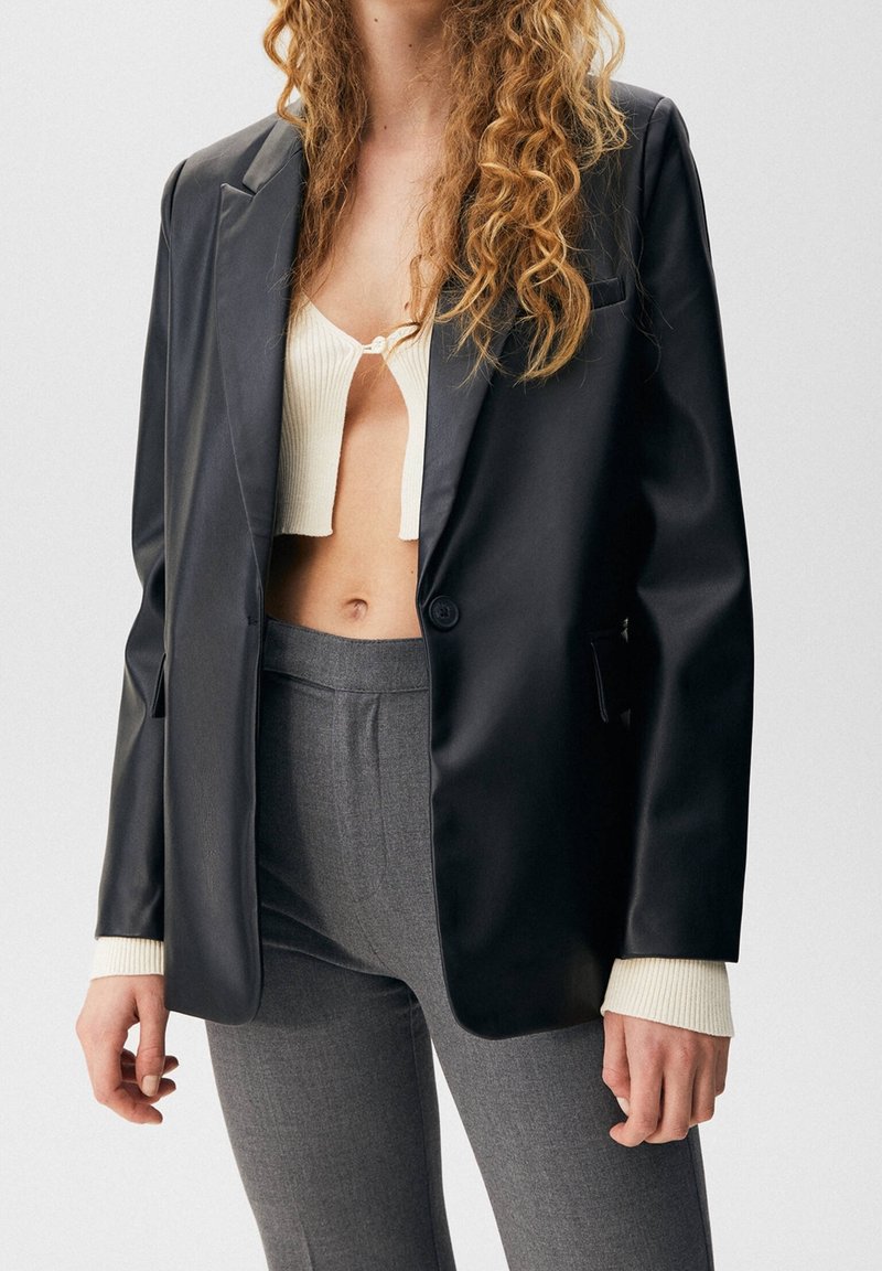 Blazer nero in ecopelle con rever a incavo, chiusura con un solo bottone e orlo curvo, abbinato a pantaloni grigi su misura e top crema a coste.