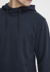 Felpa con cappuccio blu navy in tessuto texturizzato, dotata di chiusura con coulisse e tasca a marsupio. Visione ravvicinata della spalla e del collo.