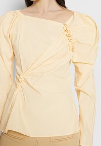 Kvinna som bär en blekgul blus med lång ärm, puffade axlar och rynkad diagonaldetalj över framsidan.