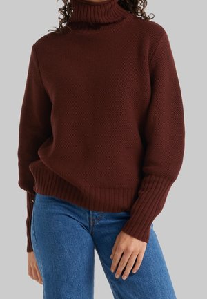 Kvinde iført en tekstureret mørkebrun rullekravesweater med ribstrikkede manchetter og kant, kombineret med blå jeans, stående mod en ensfarvet baggrund.