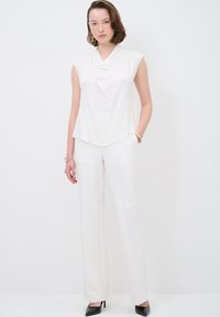 Top bianco senza maniche con scollo drappeggiato abbinato a pantaloni bianchi a gamba larga. La modella indossa scarpe con tacco nero e un bracciale.