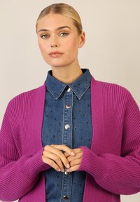 Fioletowy sweter dziergany z ryżowym wzorem, otwarty z przodu, z długimi rękawami, nałożony na niebieską koszulę dżinsową z czarnymi ćwiekami i zapinanymi na napy guzikami.